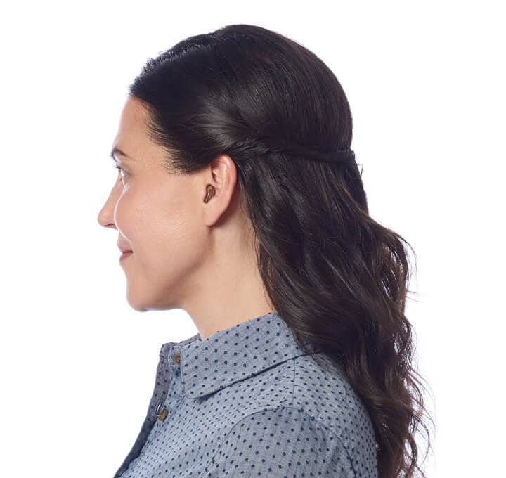 The profile of a middle-aged caucasian woman with long dark hair wearing the Starkey Genesis AI in her left ear.
