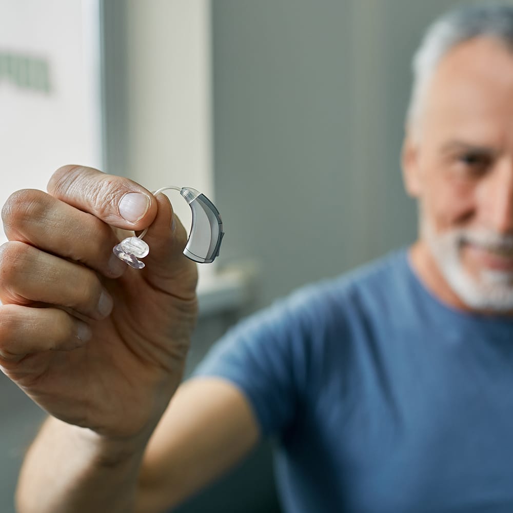 Senior man holding BTE hearing aid in hand on foreground, close-up.