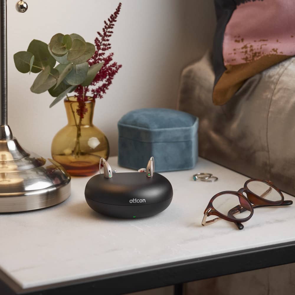 A pair of Oticon hearing aids in docking station placed on nightstand.