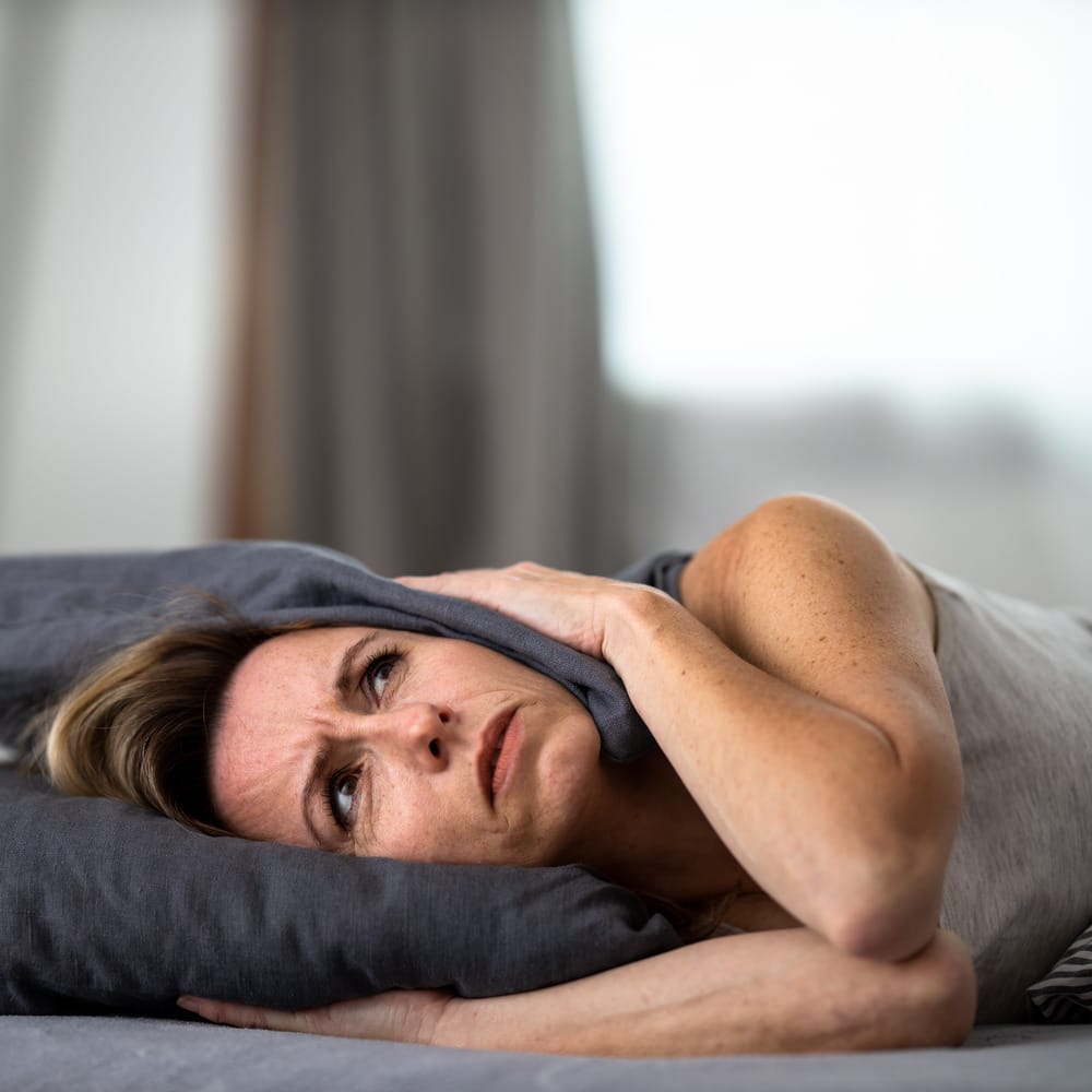 Middle-aged woman in bed, pressing both sides of pillow to head, visibly suffering from headache/tinnitus