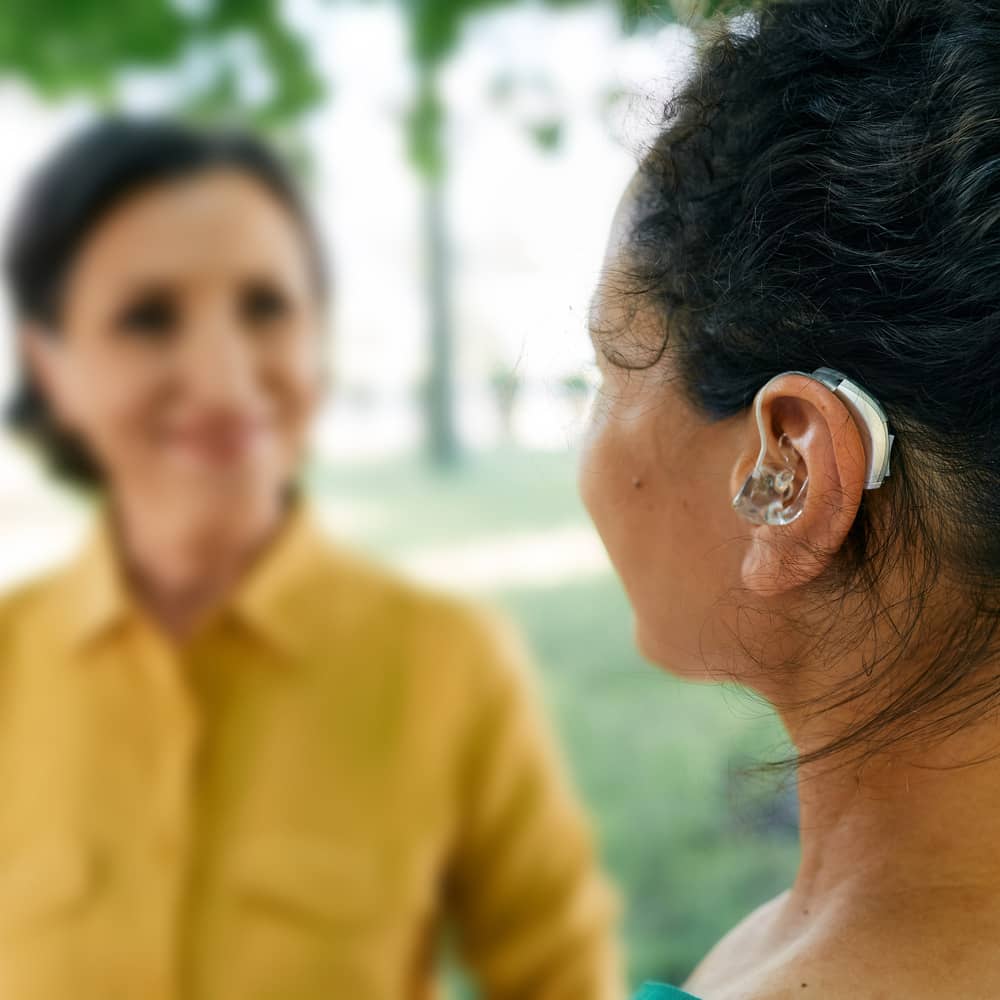 Woman wearing hearing aid speaking to friend in a park.