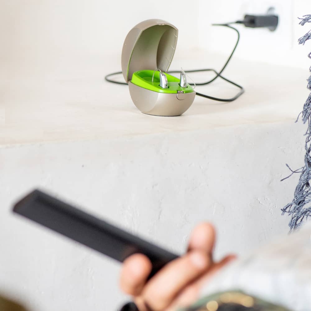 Hand holding a smartphone, Phonak hearing aids inside charger in background.