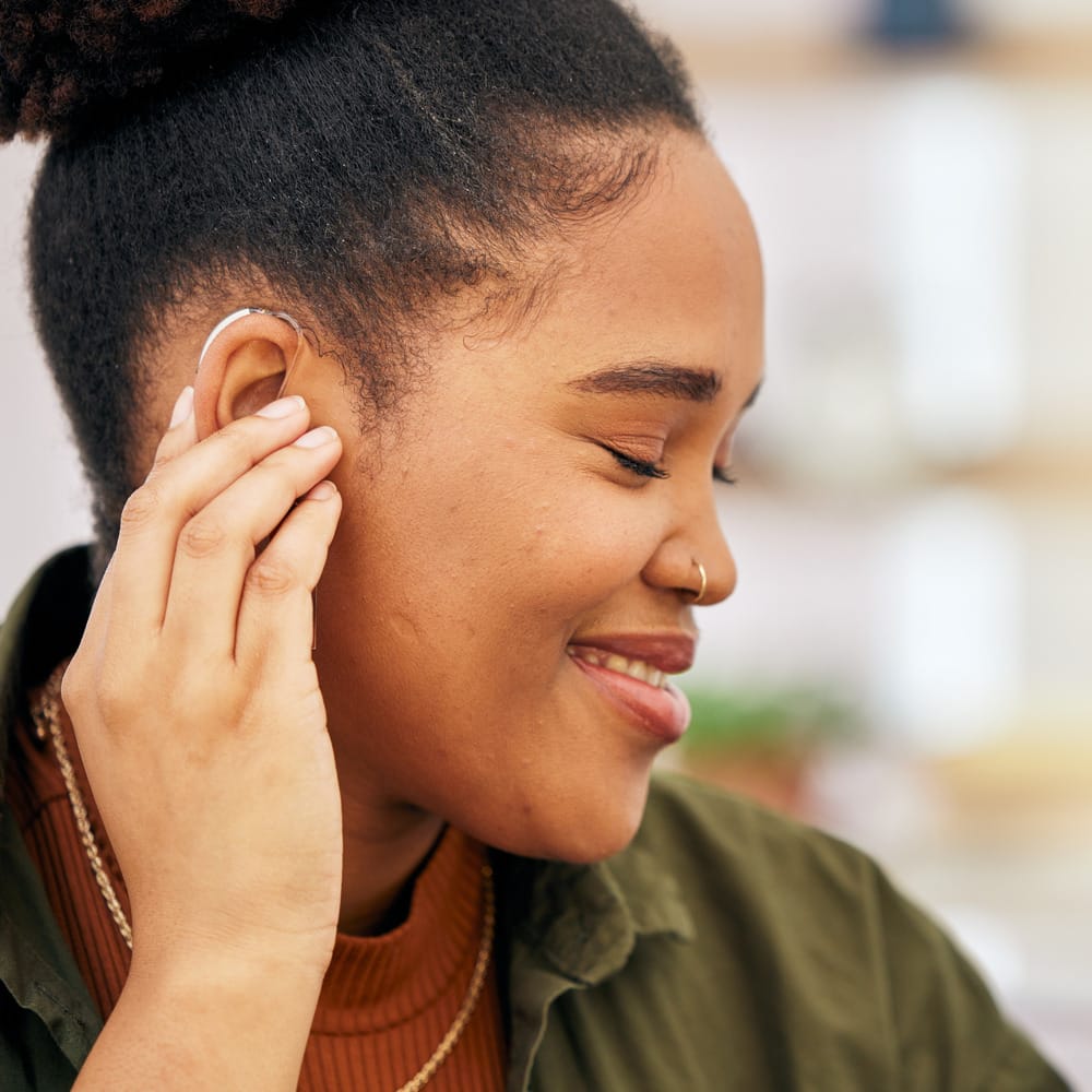 Woman inserting hearing aid into right ear, looking down, smiling.