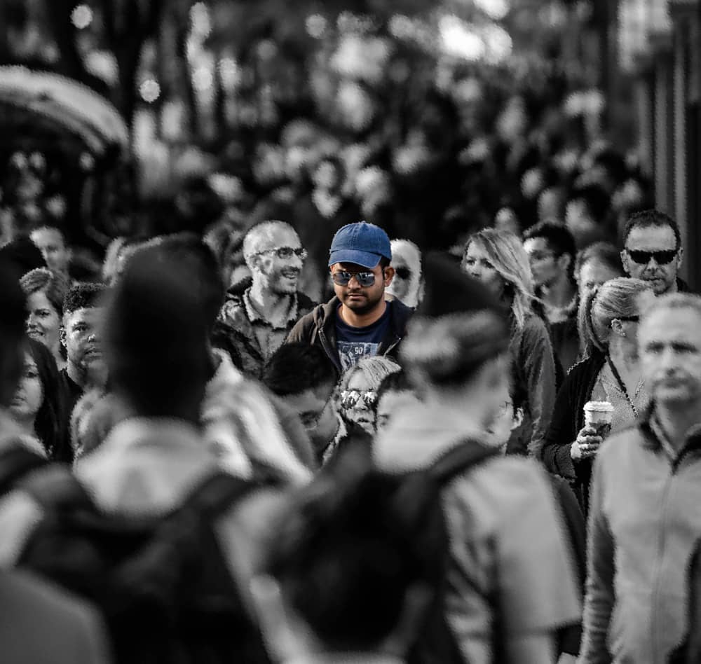 Photograph of man in glasses with blue hat in a greyscale crowd to visualise effects of hearing distortion and solitude.