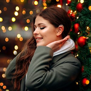Young woman with long brown hair wearing a hearing aid, standing beside a Christmas tree.