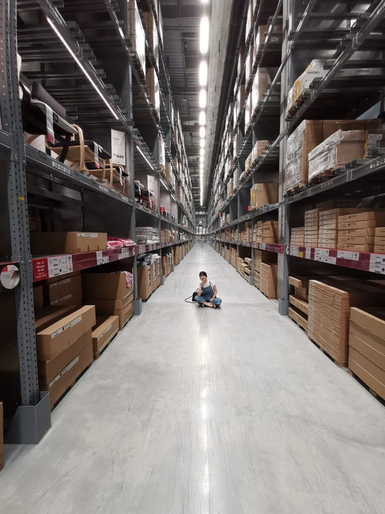 Photograph of small woman sitting cross-legged in middle of warehouse isle smiling at the camera with hand under chin.